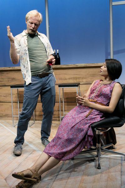 A man holding a glass gestures animatedly while standing by a bar, as a seated woman in a pink dress looks up at him, also holding a drink. The scene appears to be set on a modern stage.