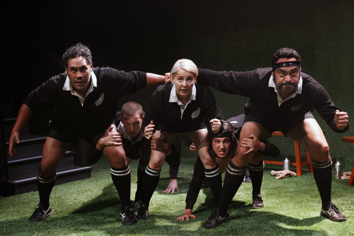 Five rugby players in black kits crouch in a formation on a grass pitch, arms linked and faces focused, preparing for a scrum under dramatic lighting.