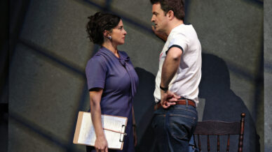 A woman in a purple dress holding a clipboard faces a man in a white T-shirt and jeans. They stand close together in an intense conversation, with dramatic lighting casting shadows on the wall behind them.