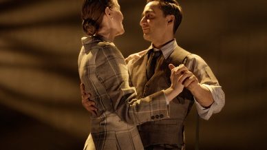 A man and woman in vintage-style clothing dance together under warm stage lighting, gazing into each other’s eyes. Shadows and a bench are visible in the background, suggesting a theatrical performance.