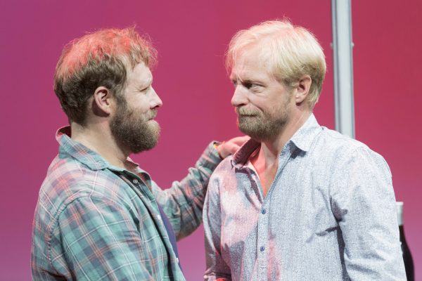 Two bearded men stand face to face on a stage with a pink background. One wears a checked shirt and has his hand on the other's shoulder; the other wears a light patterned shirt. They share a serious, intense moment.