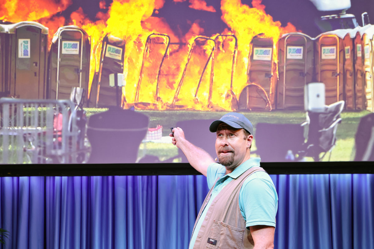A man in a blue shirt and cap stands on stage, pointing at a large screen showing portable toilets engulfed in flames. The background behind him is covered with blue curtains.