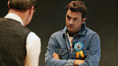 A man in a denim jacket with a blue and orange ribbon on his chest stands with arms folded, looking sceptically at another man in glasses who is seen from behind.