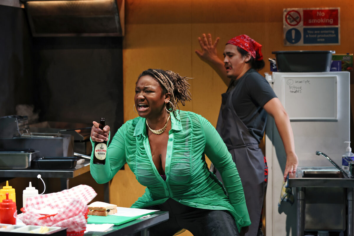 A woman in a green blouse holds a bottle of condiment and shouts in a commercial kitchen, while a person in an apron and red bandana reacts with raised hands in the background. Sandwich ingredients are on the worktop.