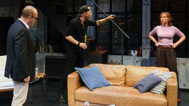 Three people are in a modern living room. A man in a black shirt and cap points aggressively at a woman with hands on hips, whilst another man in glasses watches. They stand around a tan sofa with cushions and a can of fizzy drink on the table.