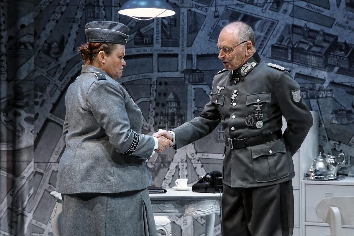 Two actors in military uniforms shake hands on stage, set against a backdrop of a vintage map. A desk with a lamp, teacup, and old-fashioned telephone is in the background. The scene suggests a formal wartime meeting.