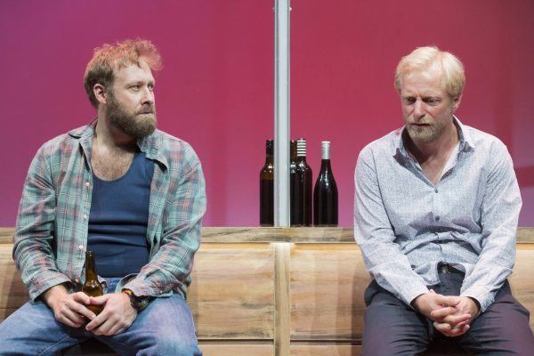 Two men sit on a bench in front of several wine bottles, looking serious. The man on the left wears a checked shirt and holds a beer, while the man on the right wears a light patterned shirt. The background is pink.
