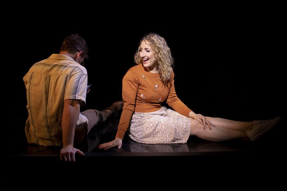 A woman with curly blonde hair, wearing a brown jumper and polka dot skirt, sits on a dark stage smiling, while a man in a short-sleeve shirt sits beside her with his back partially turned.