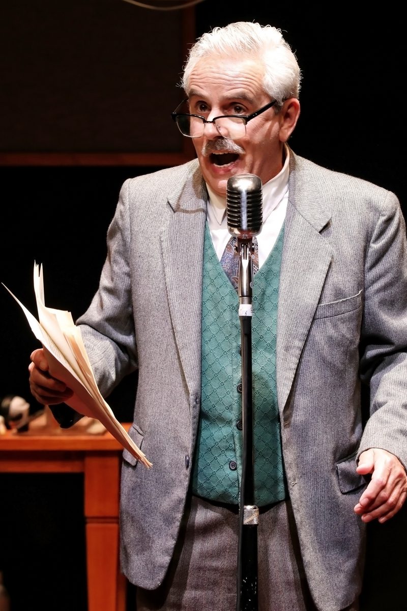 An older man with grey hair and a moustache, wearing glasses, a grey suit, and a green waistcoat, stands in front of a vintage microphone, holding papers and speaking passionately.
