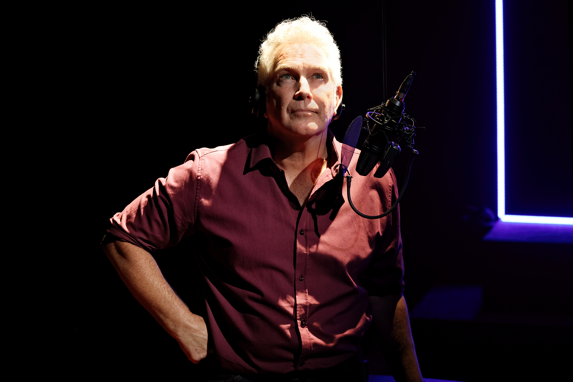 A man with grey hair wearing a maroon shirt stands in a dark room, illuminated by a spotlight. He is in front of a microphone, with one hand on his hip, looking upwards thoughtfully.
