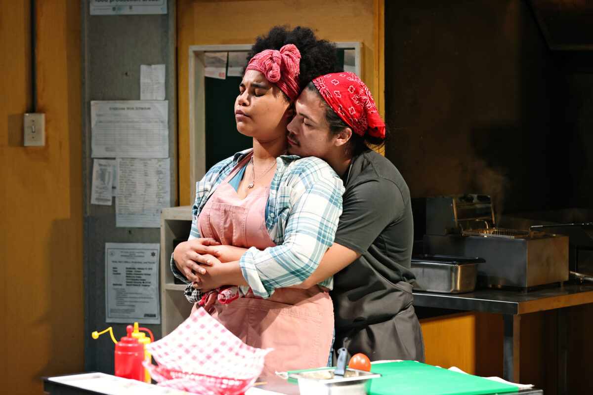 Two women stand in a kitchen. One, wearing a pink apron and headband, closes her eyes whilst the other, in a red bandana, embraces her from behind. Cooking supplies and trays are visible on the worktop.