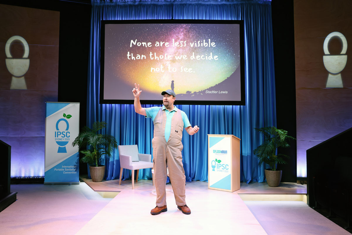 A man in overalls speaks on stage in front of a screen displaying the quote, None are less visible than those we decide not to see. The stage is decorated with plants, blue curtains, and IPSC event banners.