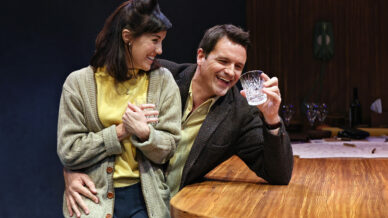 A woman sits on a man's lap, both smiling and laughing. The man holds a glass and leans on a wooden table set with crockery and a wine bottle. They appear to be enjoying a light-hearted moment together indoors.