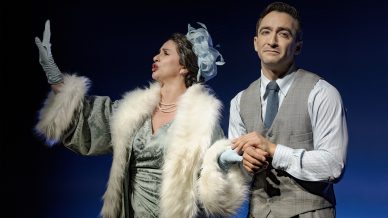 A woman in an elegant fur-trimmed dress and hat gestures dramatically, while a man in a grey waistcoat and tie stands beside her, holding her arm. The background is dark and theatrical, suggesting a stage performance.