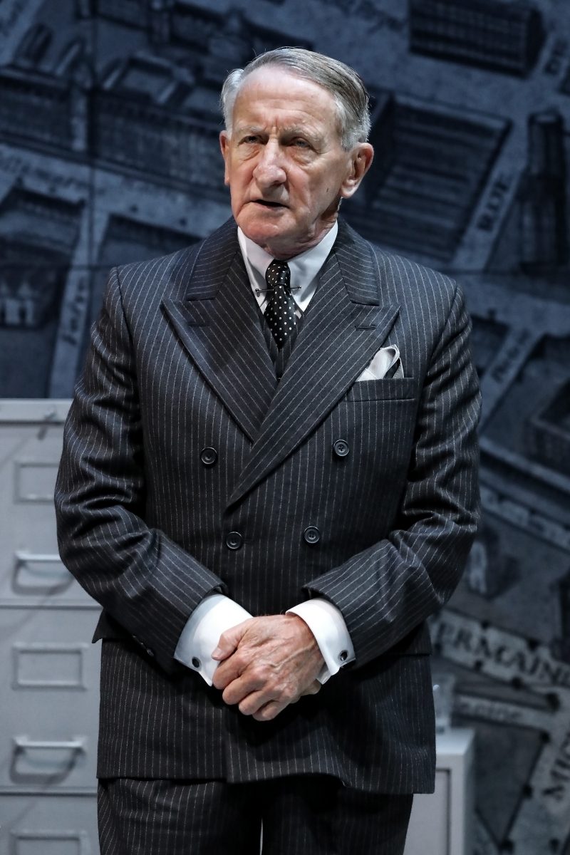 An older man in a dark pinstripe suit and polka-dot tie stands in front of a grey backdrop with filing cabinets and map-like graphics, looking serious with his hands clasped.