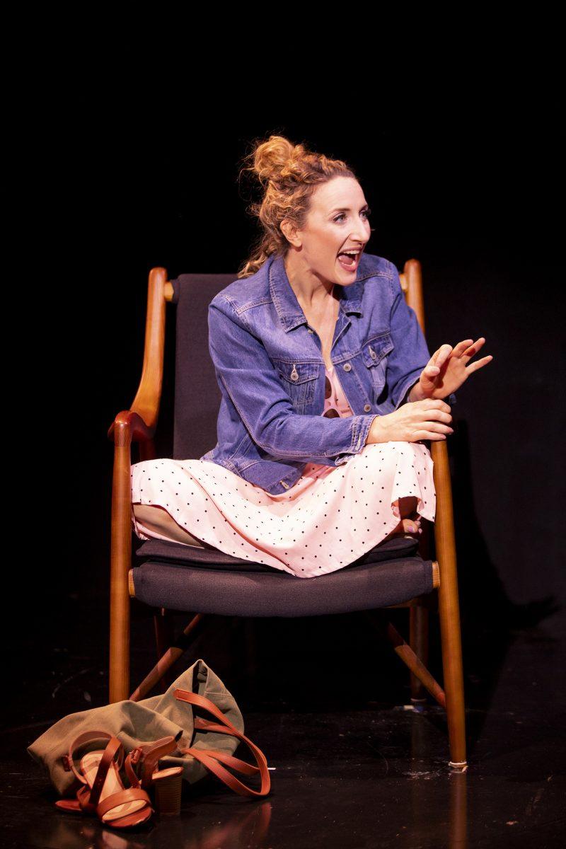 A woman with curly hair in a messy bun, wearing a denim jacket and a pink spotty dress, sits cross-legged on a chair while smiling and gesturing. A green bag and brown sandals are on the floor beside her.