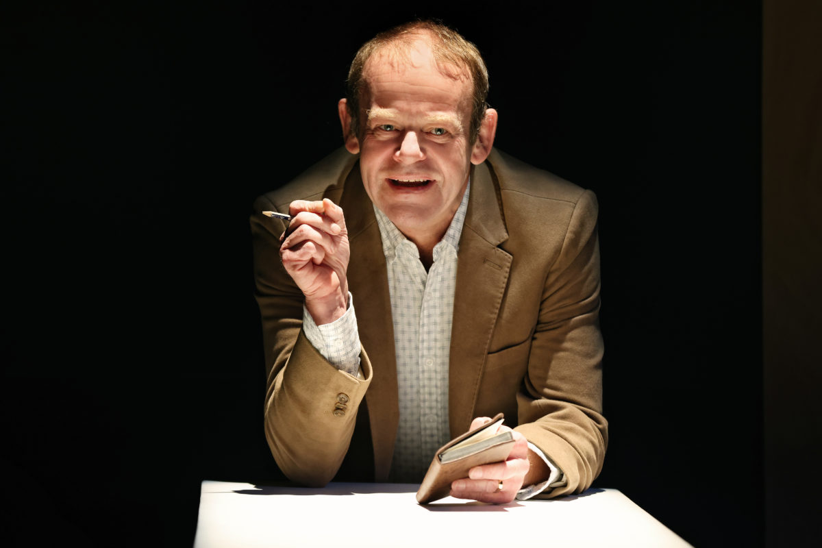 A man in a suit jacket sits at a table under a spotlight, holding a pen and notepad, and smiling directly at the camera against a dark background.