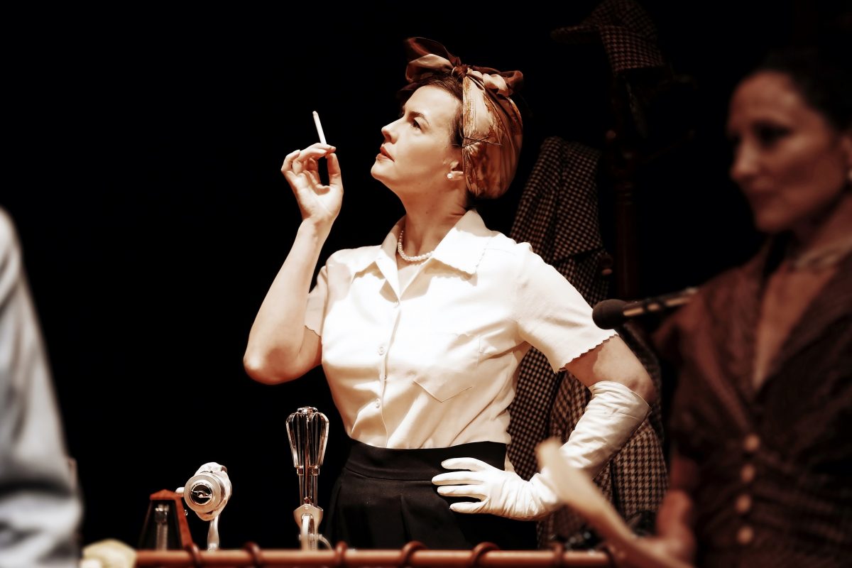 A woman in vintage clothing and gloves stands at a microphone, holding a cigarette and looking thoughtful. Another blurred figure is nearby, suggesting a dramatic or theatrical scene.