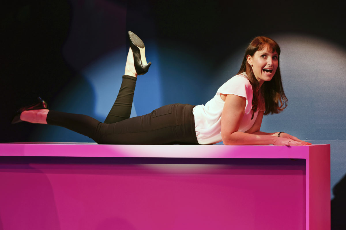 A woman in black trousers and a white blouse lies on her stomach on a bright pink table, one leg bent upward, and looks forward with a surprised or expressive facial expression under dramatic lighting.