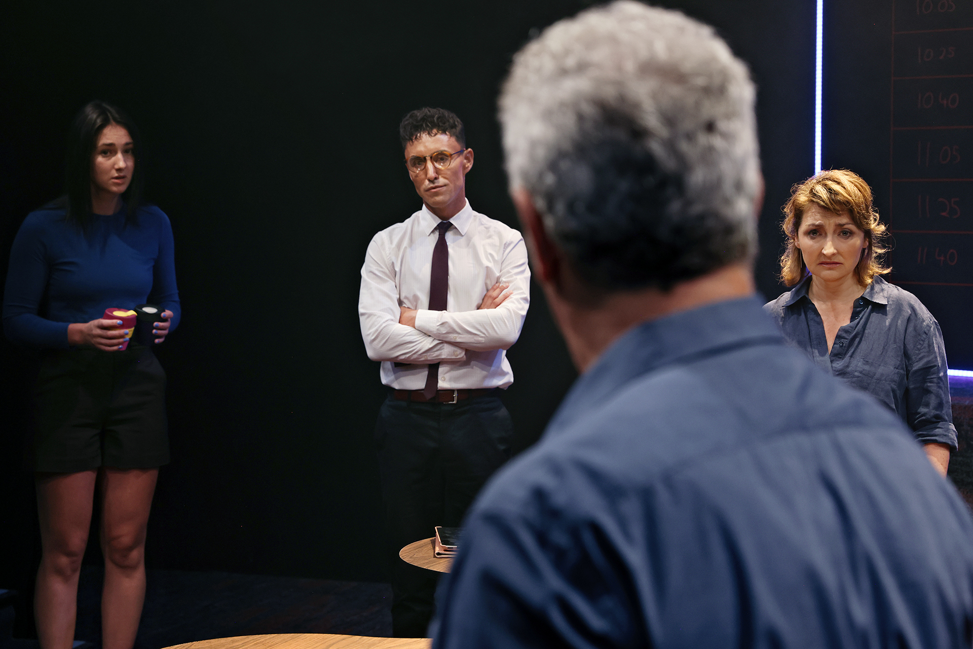 Four people stand in a dimly lit room, three facing a man with grey hair seen from behind. Two women and a man look serious and attentive, suggesting a tense or important discussion. A blackboard with times is visible in the background.