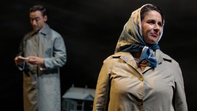 A woman in a light trench coat and patterned headscarf stands in focus, looking to the side and smiling. A man in a similar coat stands blurred in the background, holding a piece of paper. The scene appears to be on a dimly lit stage.