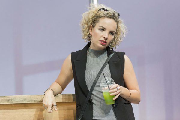 A woman with curly blonde hair leans against a wooden counter, holding a green drink in a clear plastic cup with a black straw. She is wearing a grey top, black waistcoat, and has sunglasses on her head.
