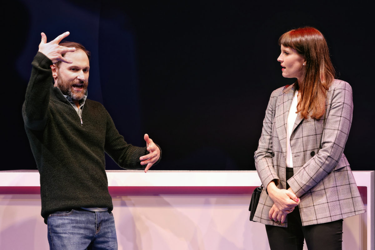A man gestures expressively with his hands while speaking to a woman in a checked blazer, who stands attentively with her hands clasped in front of her. They are indoors, in front of a dark background.