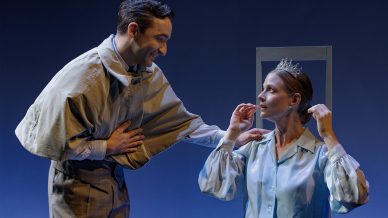 A man in vintage clothing smiles and gestures towards a seated woman wearing a crown. The woman adjusts the crown on her head, and both are dressed in formal, old-fashioned attire against a blue background.