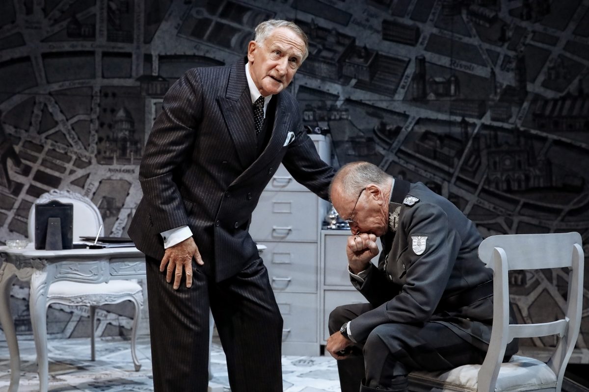 Two older men in suits are on stage; one stands, looking serious, while the other, dressed in a military uniform, sits and rests his head on his hand, appearing distressed. The set has a desk and a wall covered with maps.