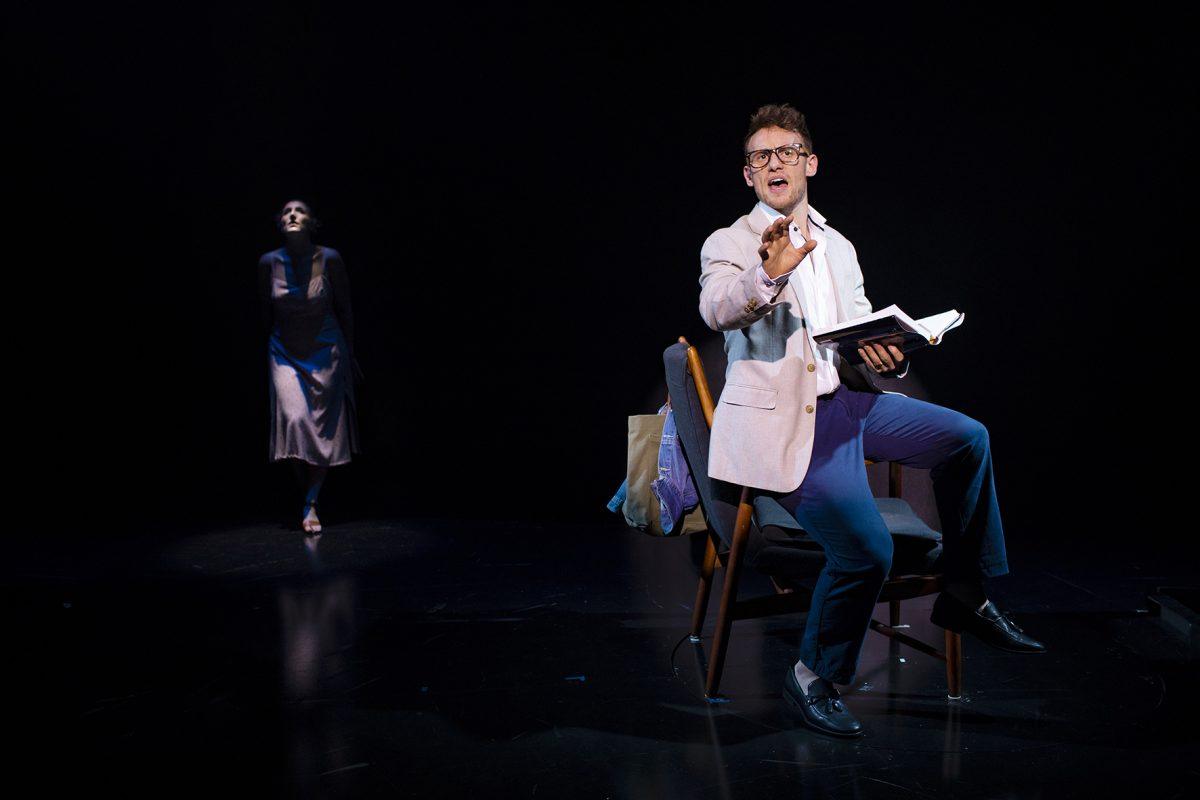 A man in glasses sits on a chair holding an open book, gesturing passionately, whilst a woman in a dress stands in the dark background, partially illuminated. The scene appears to be from a theatrical performance.
