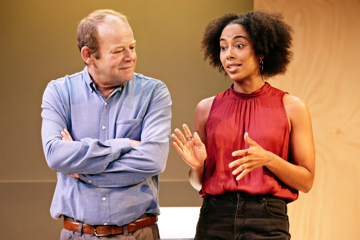 A man in a blue shirt stands with his arms folded, smiling at a woman in a sleeveless red top who is gesticulating with her hands while talking. Both appear to be on a stage with a neutral background.