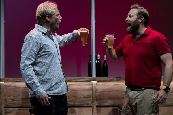 Two men face each other, holding beer glasses in a toast and smiling. One wears a patterned shirt and the other wears a red polo and khaki trousers. Three wine bottles are visible in the background, set against a pink wall.