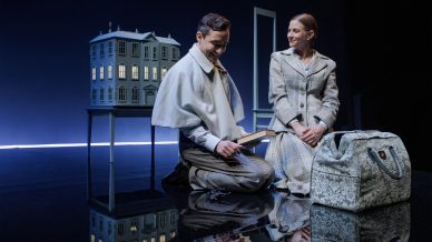 Two actors in period clothing kneel onstage beside a miniature house on a table. The man is reading a book and smiling; the woman looks at him, smiling. A patterned bag sits on the reflective floor in front of them.