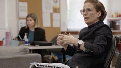 A woman with glasses gestures while speaking in an office, holding a notebook on her lap. Another woman works at a desk in the background. The room includes a noticeboard and office equipment.