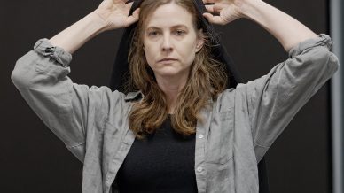A woman with long brown hair, wearing a grey button-up shirt over a black top and dark jeans, stands indoors adjusting a black cloth or veil on her head with both hands. A chair with papers is visible in the background.