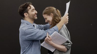 A man and woman stand facing each other, smiling and laughing. The man holds up a sheet of paper, while the woman holds a script, both appearing joyful against a plain dark background.
