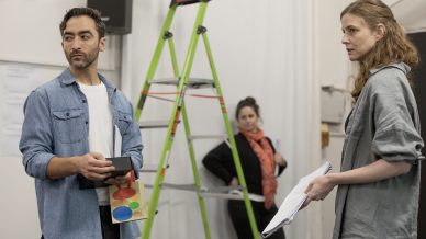 A man holding a colourful gift bag and box stands next to a woman with papers; another woman stands in the background by a green ladder in a room with white walls.