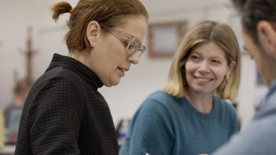 Two women sit together in conversation; one with glasses and a black jumper is writing, while the other with blonde hair and a blue jumper smiles at her. A blurred person is in the foreground.
