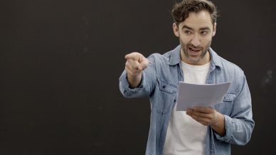 A man with wavy hair wearing a denim shirt and white T-shirt holds a script in one hand and points forward with the other, appearing to speak or perform in front of a plain dark background.