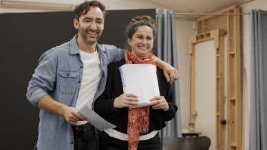 Two people stand side by side, smiling. The man has his arm round the woman. Both hold papers, suggesting they are at a rehearsal. The background features curtains and a wooden frame, creating a casual, indoor setting.