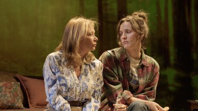 Two women sit closely on a wooden bench in a room with forest scenery in the background. One wears a patterned blouse and khaki trousers, and the other wears a checked shirt and jeans. They appear to be having a serious conversation.