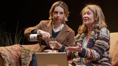 Two women sit on a sofa. One pours wine into a glass while the other gestures as she speaks. A Toshiba laptop sits in front of them. They appear engaged in conversation in a cosy setting.