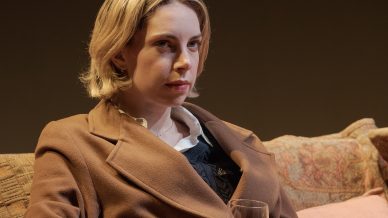 A woman with short blonde hair sits on a patterned sofa, wearing a brown coat and holding a glass of water, looking thoughtful.