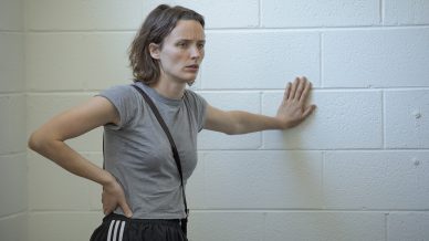 A person with short brown hair, wearing a grey t-shirt and black trousers with white stripes, stands indoors, leaning one hand against a white brick wall and resting the other on their hip, looking concerned.