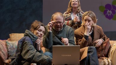 Four people sit closely together on a sofa, focused on a Toshiba laptop. Two women look concerned, an older man adjusts his glasses, and a woman at the back stands, appearing anxious. They are indoors with cosy decor.