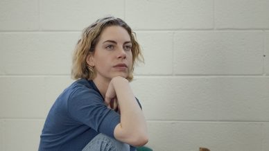 A person with short, wavy hair sits on the floor against a white brick wall, resting one arm on their knee and looking thoughtful. They wear a blue top, light jeans, and boots. Bags and other items are nearby.