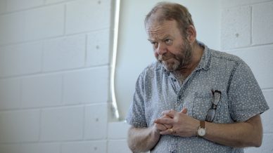 A middle-aged man with a beard, wearing a patterned short-sleeve shirt and wristwatch, stands indoors with clasped hands and spectacles hanging from his shirt, looking slightly downward with a thoughtful expression.