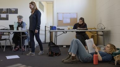 Four women are in a casual meeting room. Two sit at a table, one stands smiling, and one reclines on the floor reading a notebook. Papers, bags, and a water bottle are scattered around them.