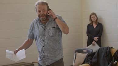 A man stands talking on the phone while holding a piece of paper, looking concerned. A woman stands in the background near a wall, watching quietly. Several chairs and a jacket are visible in the room.