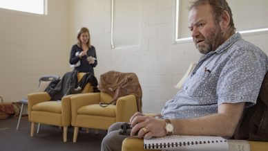 A man sits in a yellow armchair holding a spiral-bound notebook, looking thoughtful. In the background, a woman stands near another armchair with a bag and jacket laid over it, in a light-filled room.